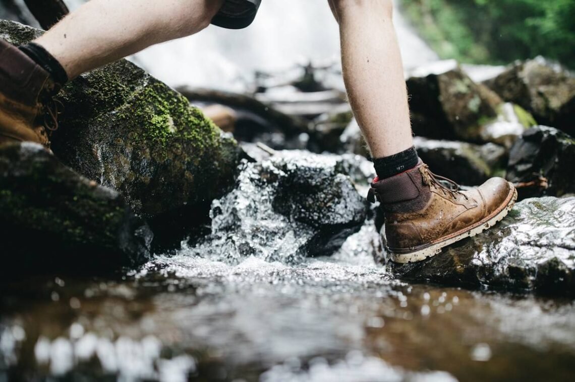 How to prevent blisters while hiking: a hiker in leather boots stepping across wet mossy rocks at a stream crossing